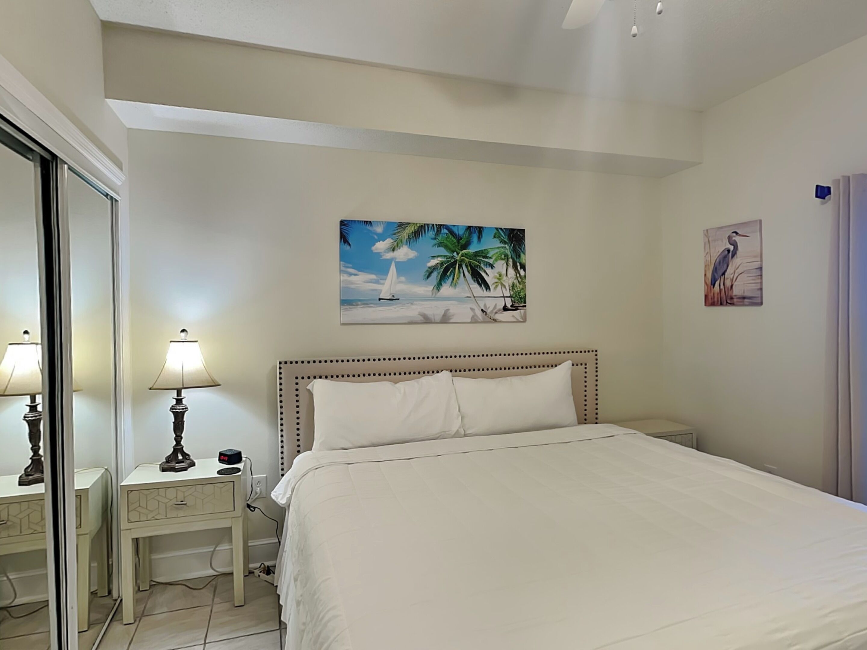 Un dormitorio con cama, una mesita de noche, una lámpara, un cuadro con escena de playa y un cuadro colgado en la pared con la imagen de un pájaro.