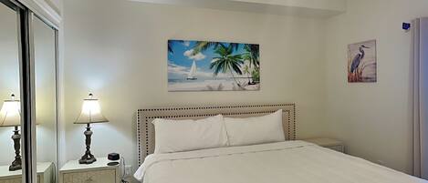 Un dormitorio con cama, una mesita de noche, una lámpara, un cuadro con escena de playa y un cuadro colgado en la pared con la imagen de un pájaro.