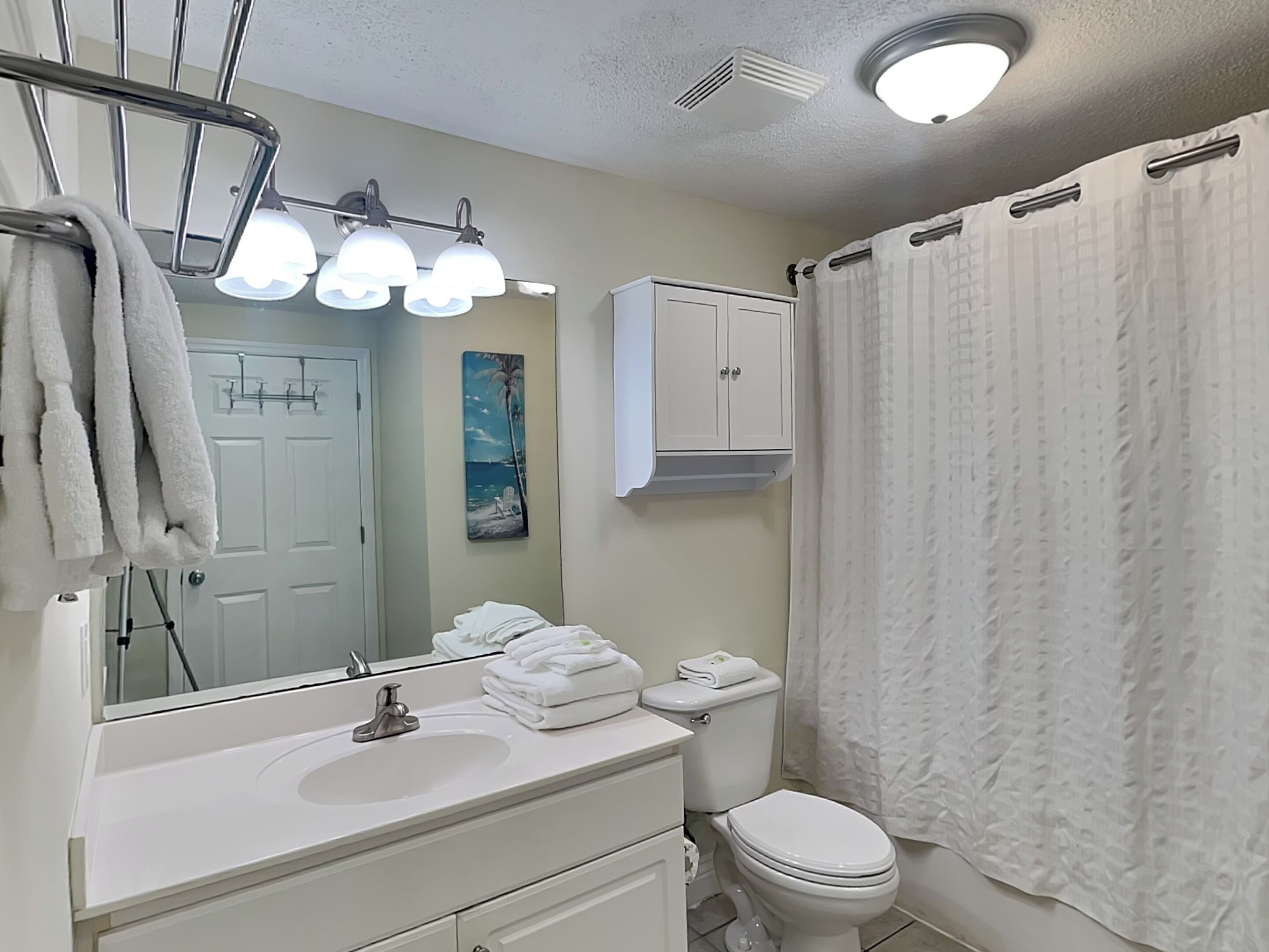 Une salle de bain avec un lavabo blanc, des toilettes et un rideau de douche. On y trouve un porte-serviettes avec des serviettes et un tableau accroché au mur.