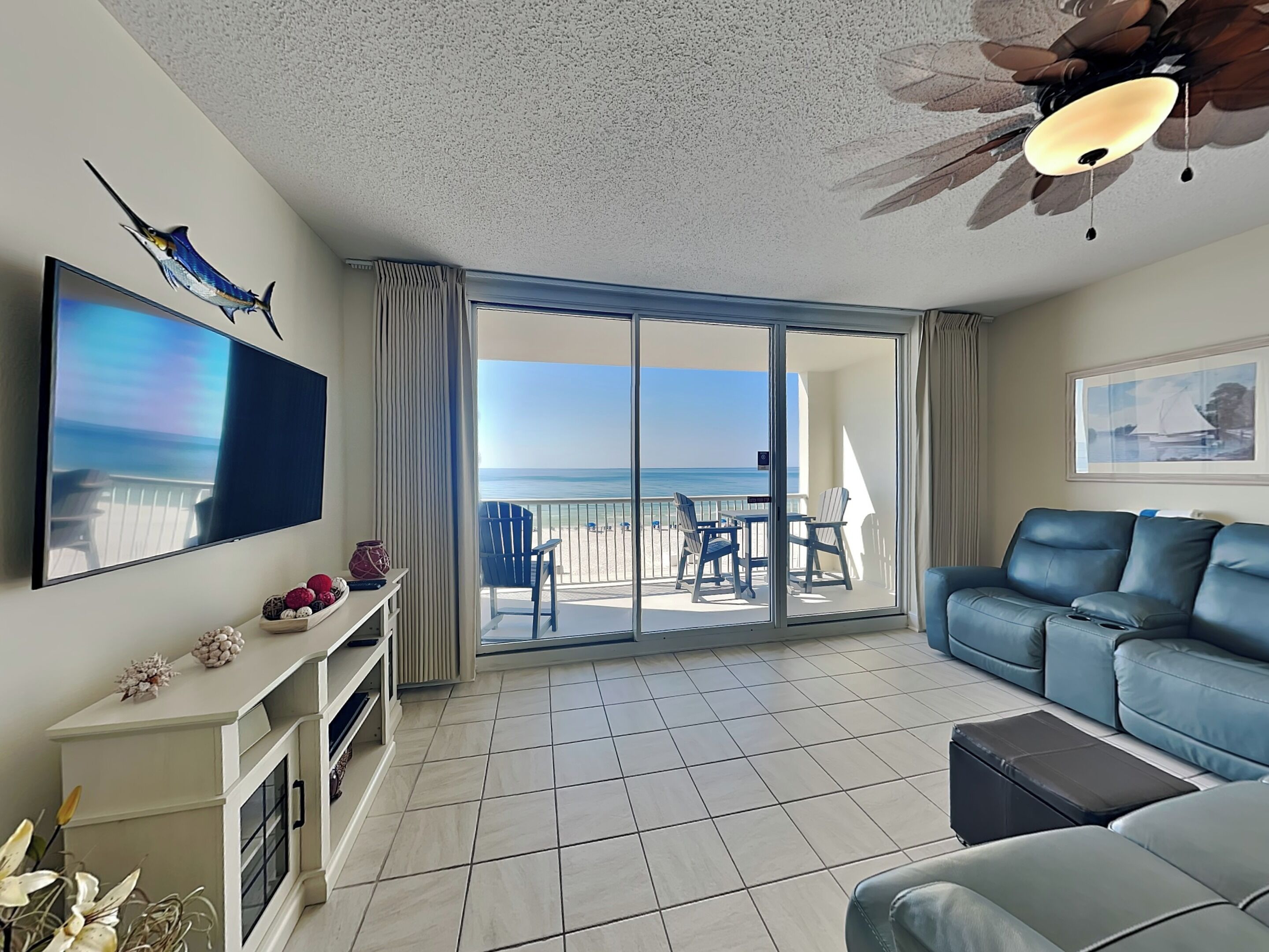 A spacious living room with a large flat-screen TV, a ceiling fan, and a balcony with a view of the beach.