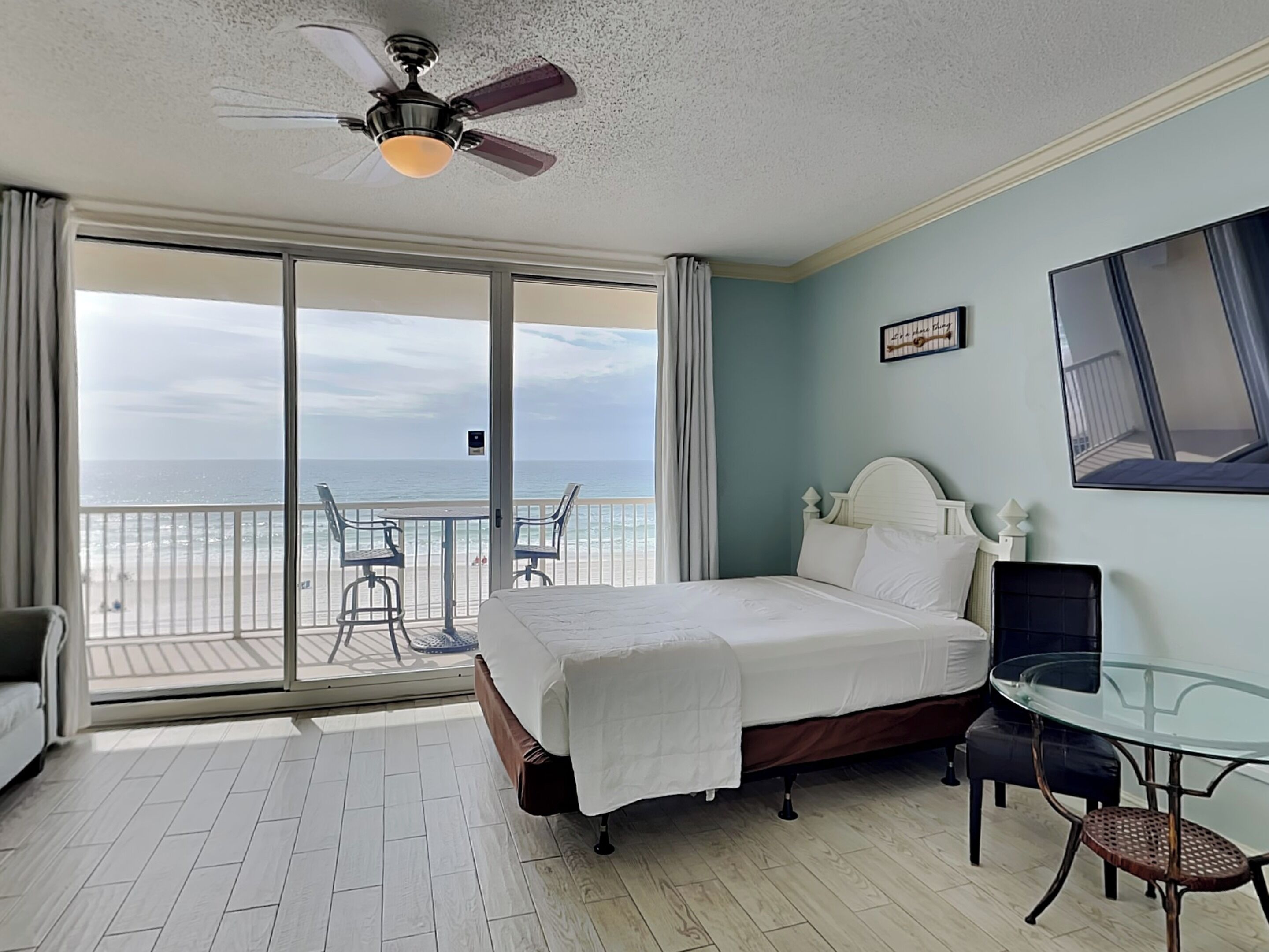 A hotel room with a bed, a glass table, a chair, a ceiling fan, and a view of the ocean.