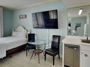 Une chambre d’hôtel comprenant un lit, une table en verre avec deux chaises, un téléviseur fixé au mur, un miroir et un lavabo avec un lave-vaisselle.