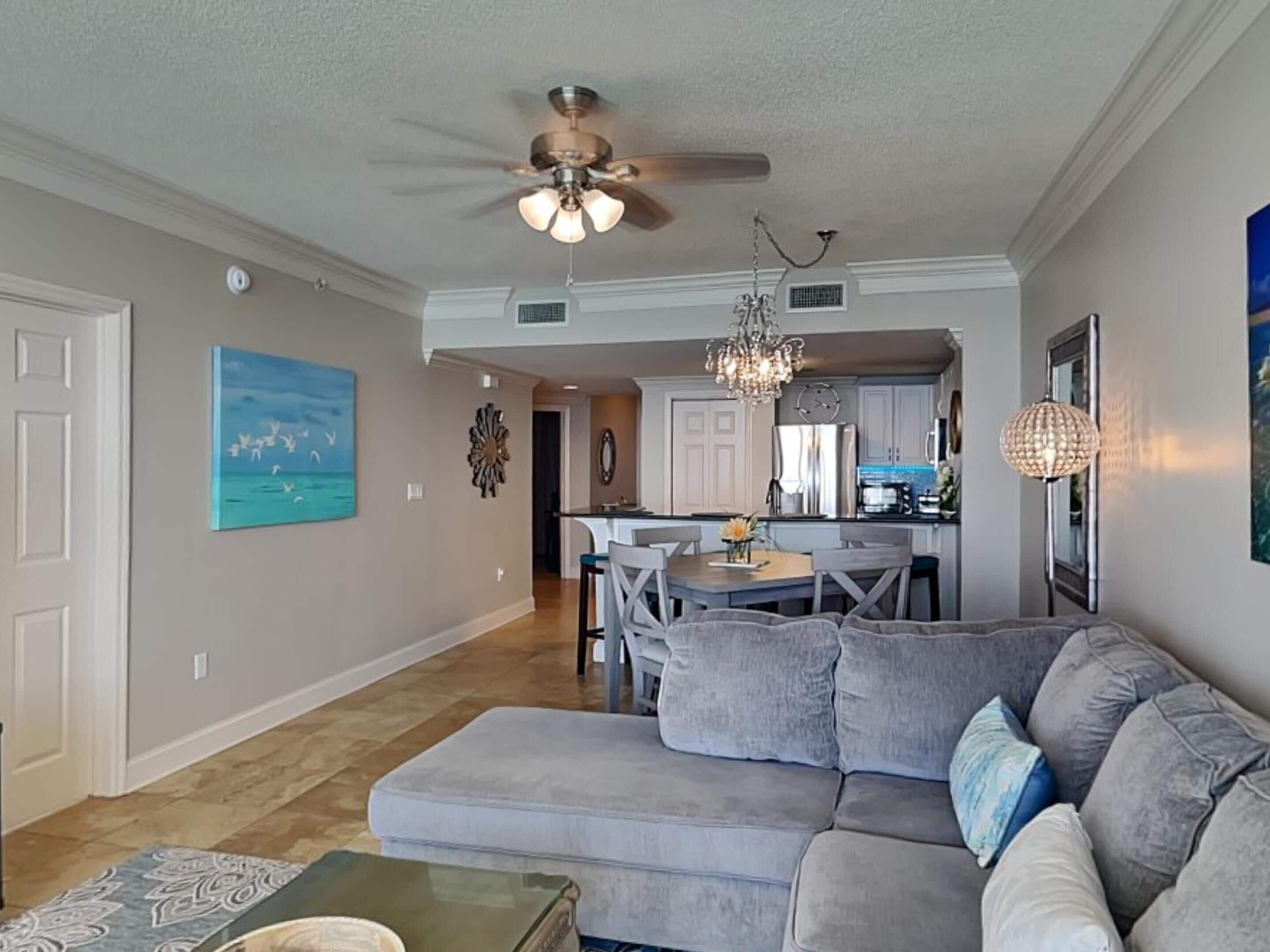 A living room with a gray sofa, a coffee table, and a chandelier. There is a dining area with a table and chairs, and a kitchen in the background.