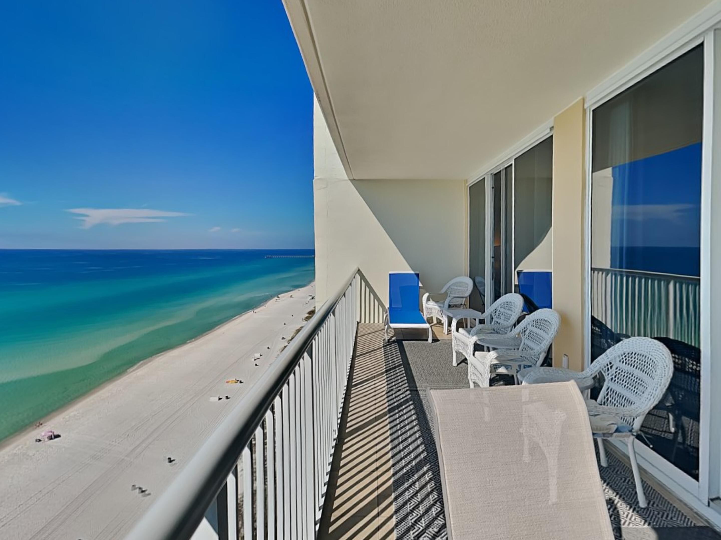 A balcony with a view of the beach and ocean, white wicker chairs, and a table.