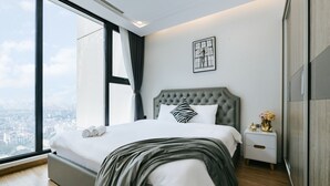 Ein modernes Schlafzimmer mit einem großen Bett, einem Nachttisch, einem gerahmten Kunstwerk und Blick auf die Stadt durch ein großes Fenster.