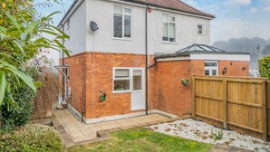 A two-story house with a brick exterior and a white upper section, featuring a front door, windows, and a small garden area with a wooden fence. - Sea Whispers Cottage (Poole)