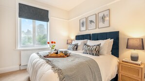 A bedroom with a large bed, a bedside table, a window with a view, and framed artwork on the wall. - Sea Whispers Cottage (Poole)