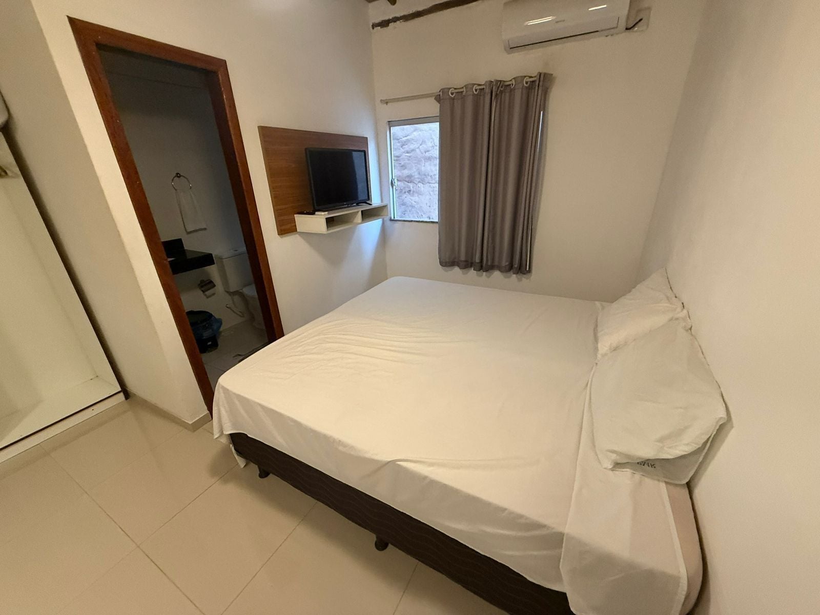 A bedroom with a bed, a TV on a shelf, a window with curtains, and a door leading to a bathroom.