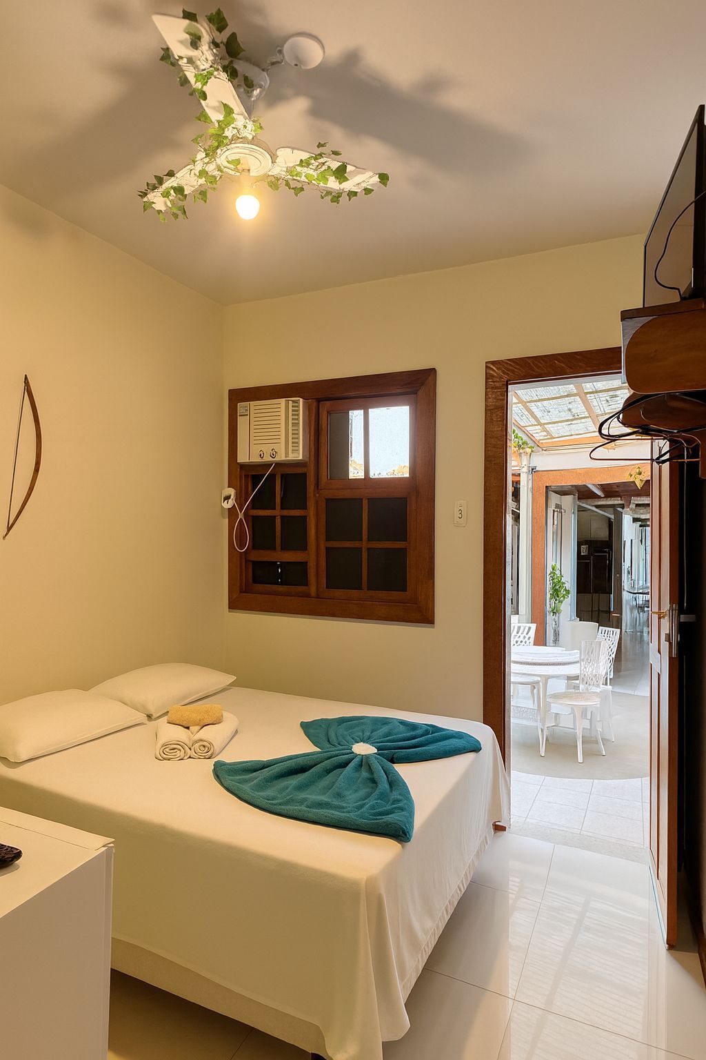 A hotel room with a bed, a ceiling fan with greenery, a window with blinds, and a view of the dining area.