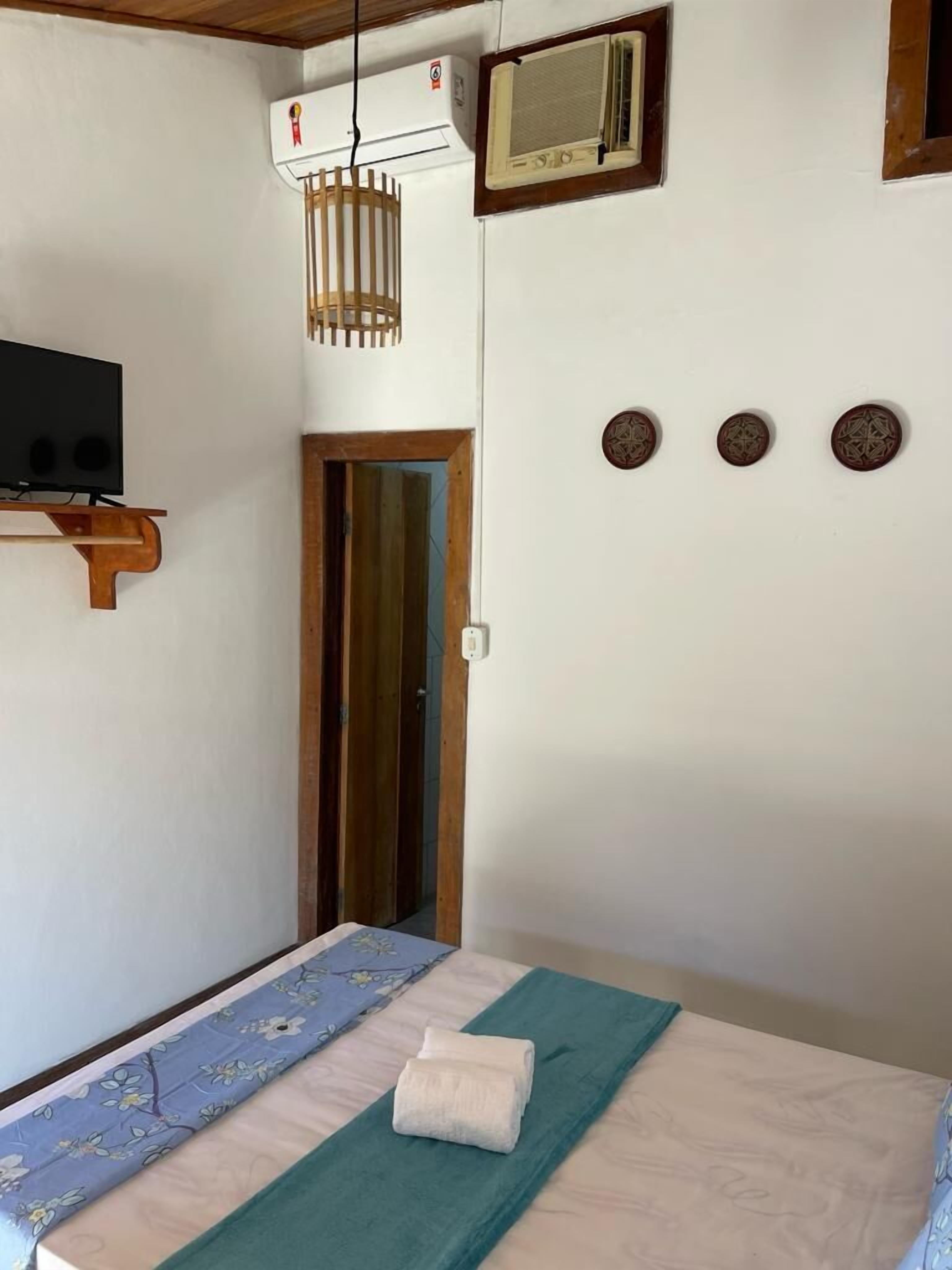 A room with a bed, a television on a wooden shelf, an air conditioning unit, a window with a wooden frame, and a door.