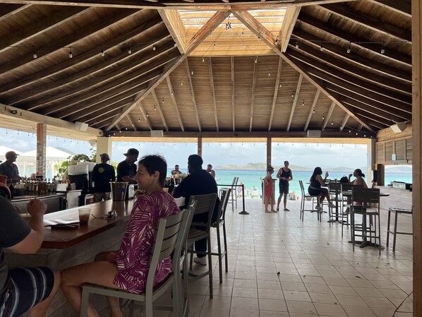 A covered outdoor dining area with wooden beams and string lights, overlooking a beach and ocean.