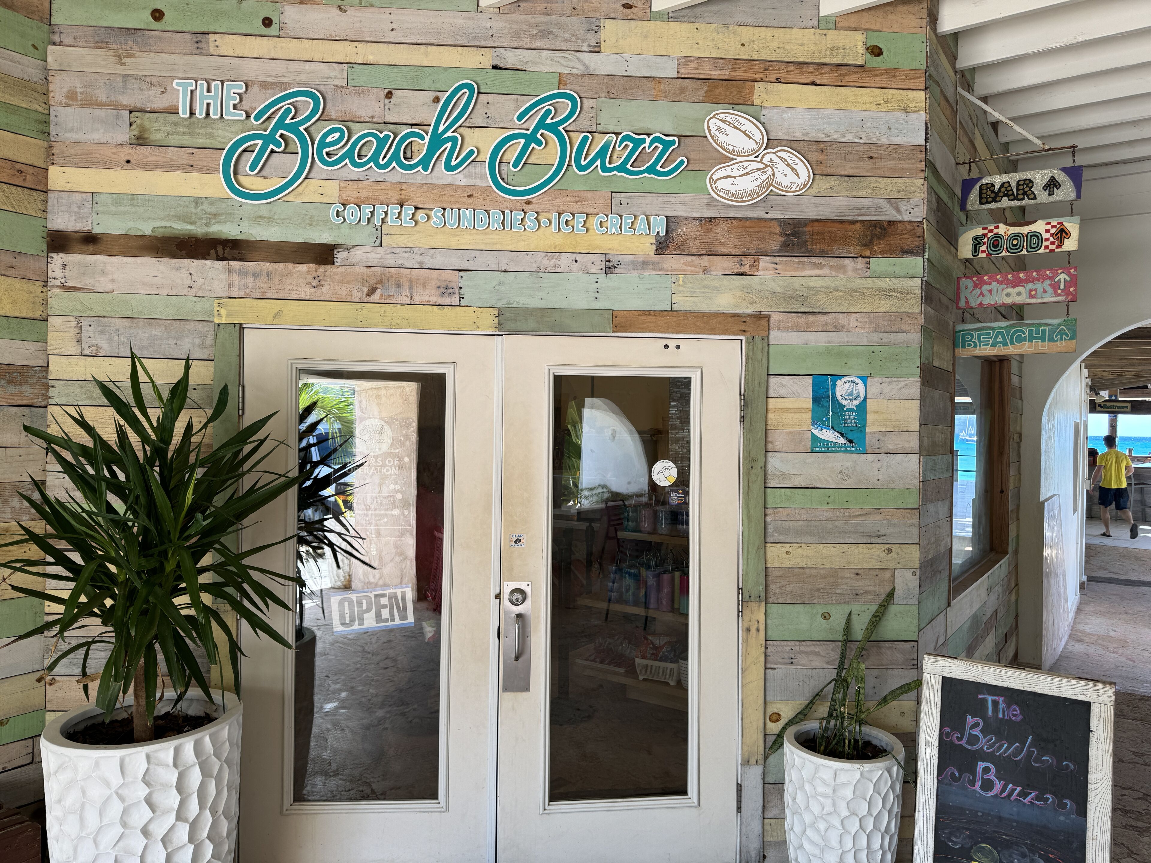 A wooden exterior wall with a sign reading "The Beach Buzz" and a chalkboard sign advertising coffee, sundries, and ice cream.