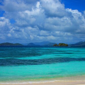 A beach with clear turquoise waters, distant islands, and a cloudy sky.