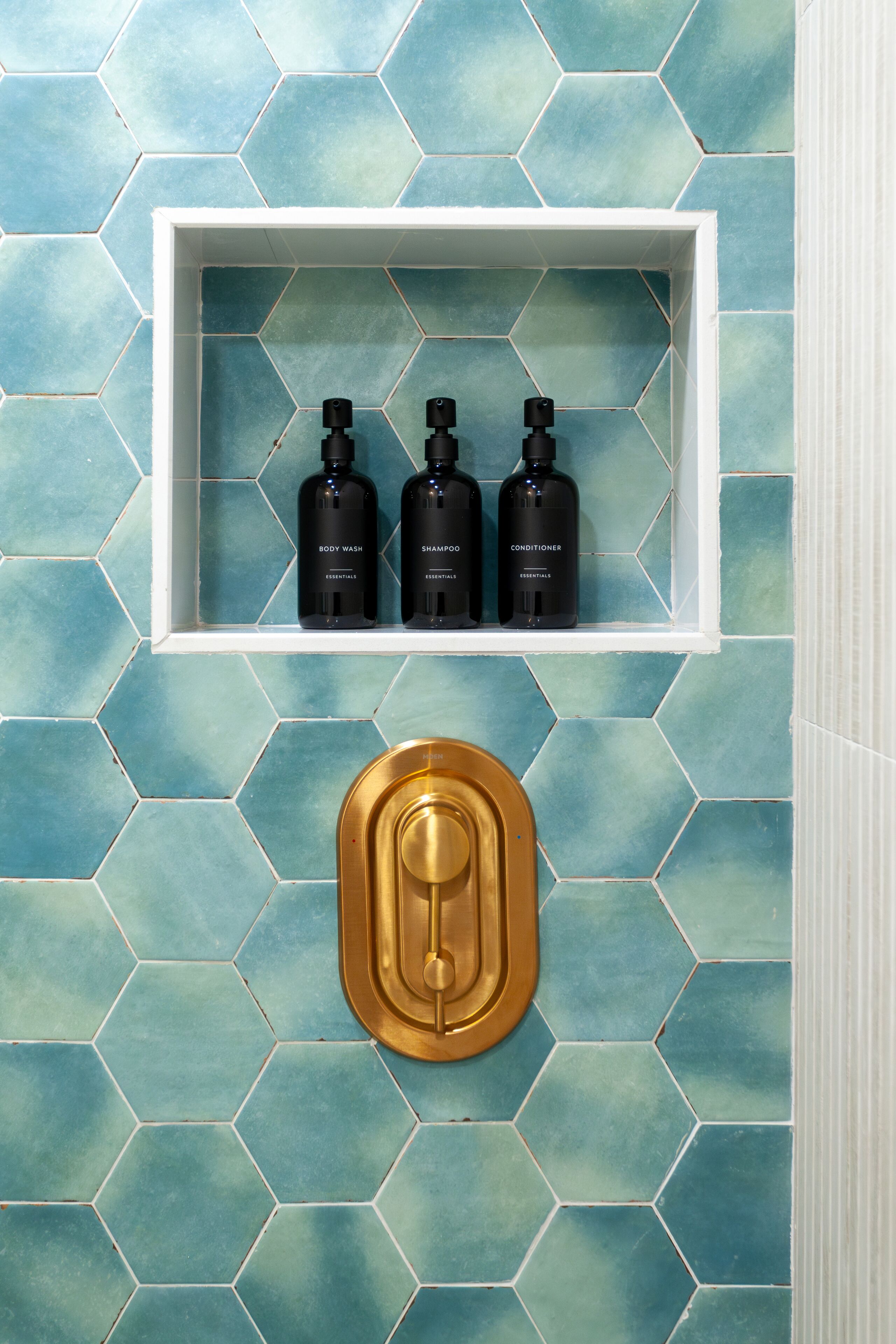 A bathroom with a hexagonal tile backsplash and a white shelf holding three black bottles labeled "BODY WASH," "SHAMPOO," and "CONDITIONER."
