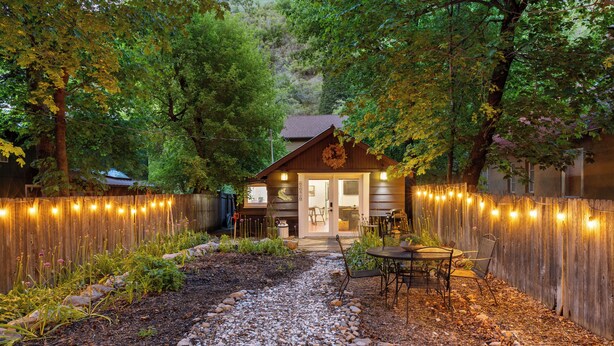 Une cabane en bois avec un chemin de gravier menant à une table et des chaises, le tout entouré d’une clôture en bois et illuminé de guirlandes lumineuses.