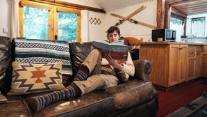 Une personne est assise sur un canapé en cuir, lisant un livre dans une pièce aménagée dans un style rustique, avec des poutres en bois, une table en bois et une fenêtre ornée de rideaux.