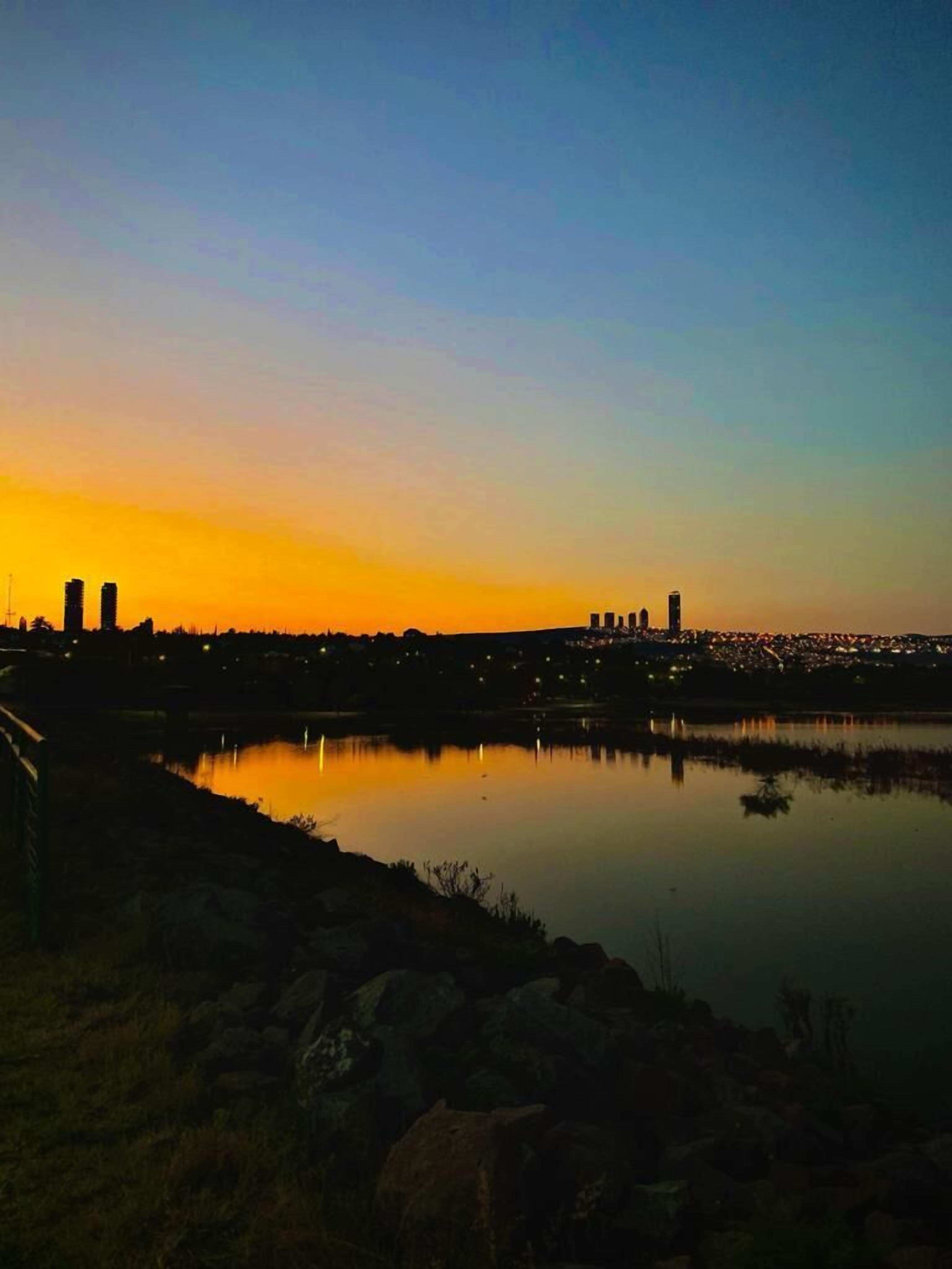 Un horizonte urbano al anochecer, con edificios silueteados contra un cielo de atardecer, una masa de agua tranquila que refleja las luces, y una costa rocosa en primer plano.