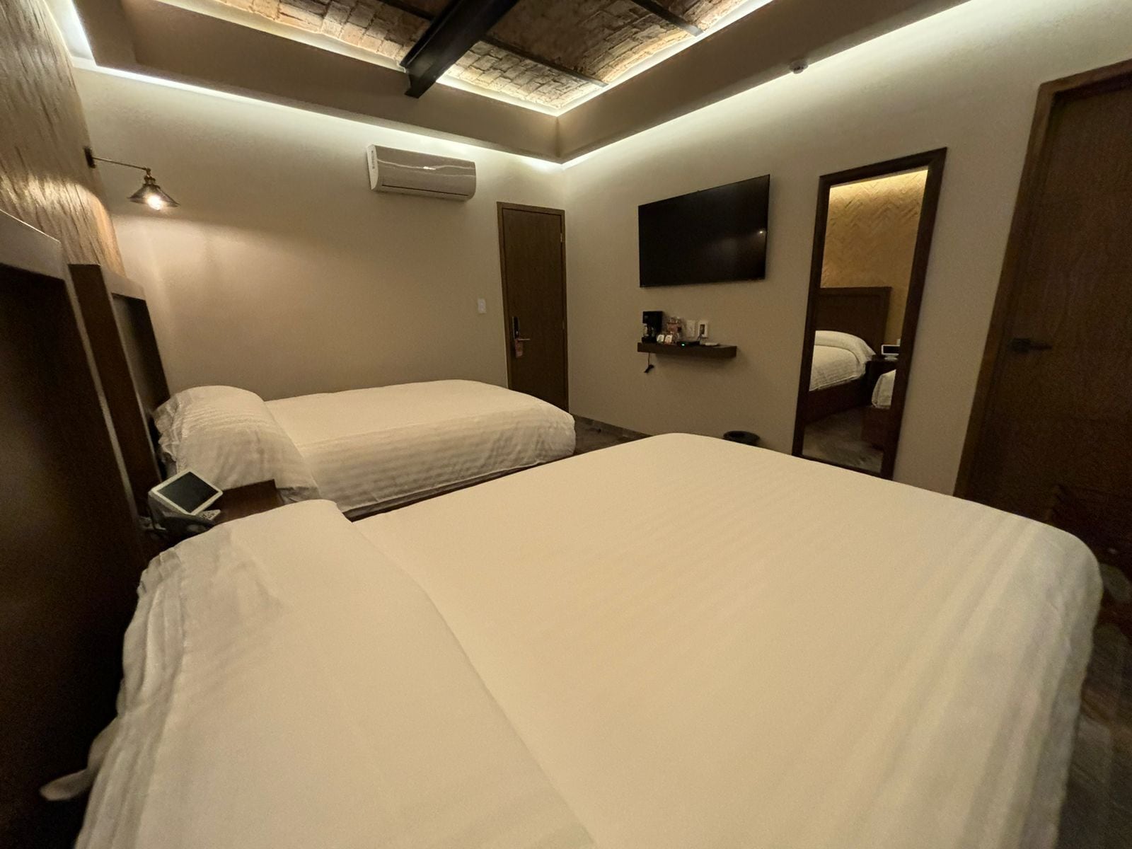 A hotel room with two beds, a flat-screen TV mounted on the wall, an air conditioning unit, and a small shelf with toiletries.