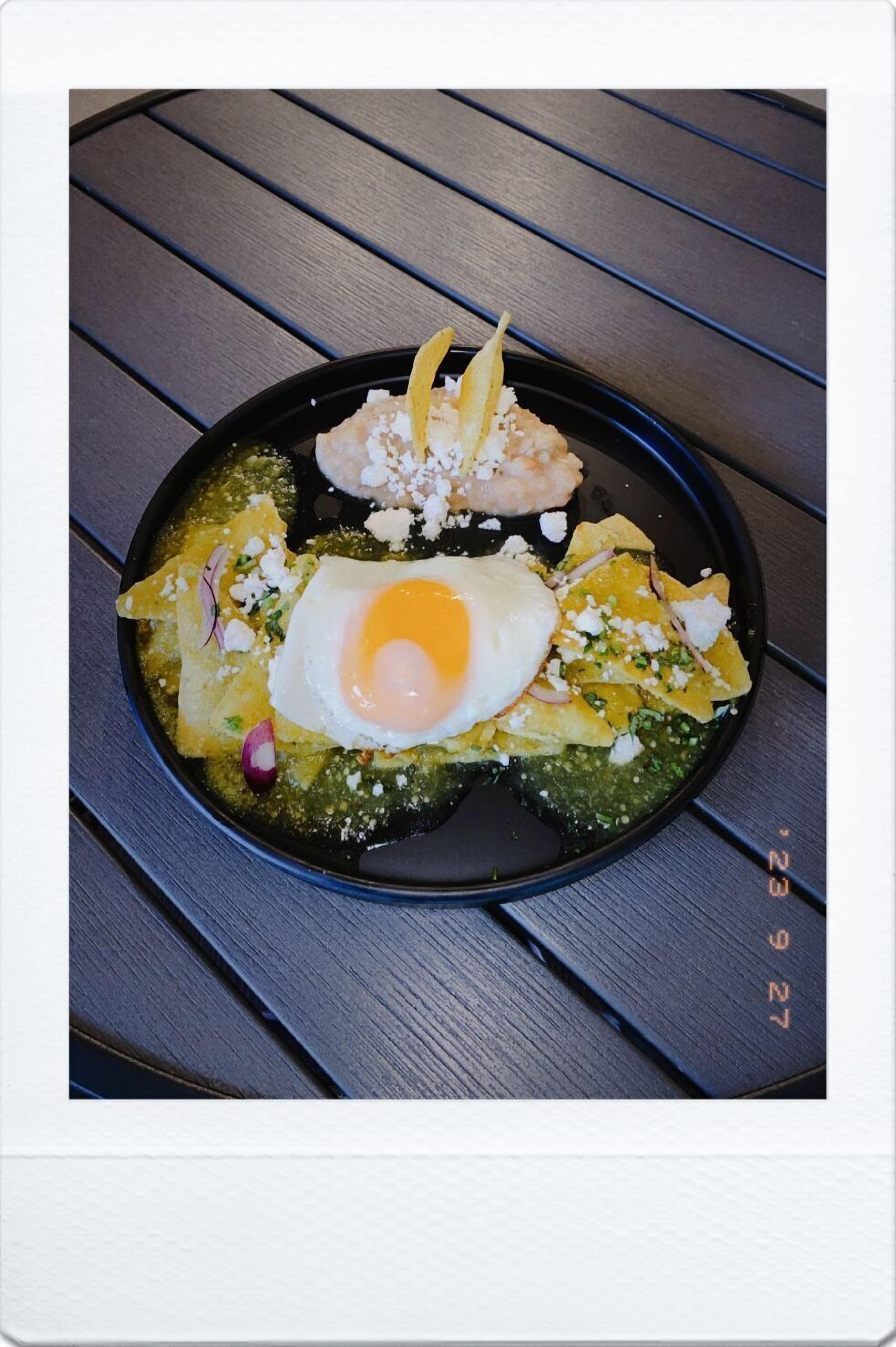 Un plato negro con un huevo frito, salsa verde y queso desmenuzado sobre una mesa de madera.