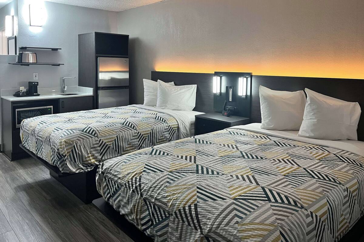 A hotel room with two beds, a kitchenette, and a nightstand with a clock.