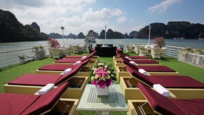 A deck with sun loungers and a table with a floral centerpiece, overlooking a body of water with boats and rocky cliffs. - Lotus D'Orient Cruise (Tuan Chau)