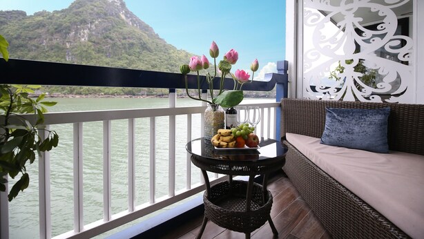 A balcony with a wicker sofa, a table with fruit and a vase of flowers, and a view of a mountain and water. - Lotus D'Orient Cruise (Tuan Chau)