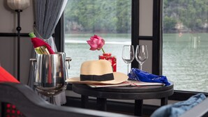 A table with a drink, a hat, a rose, and a glass, set against a window with a view of a lake and forest. - Lotus D'Orient Cruise (Tuan Chau)