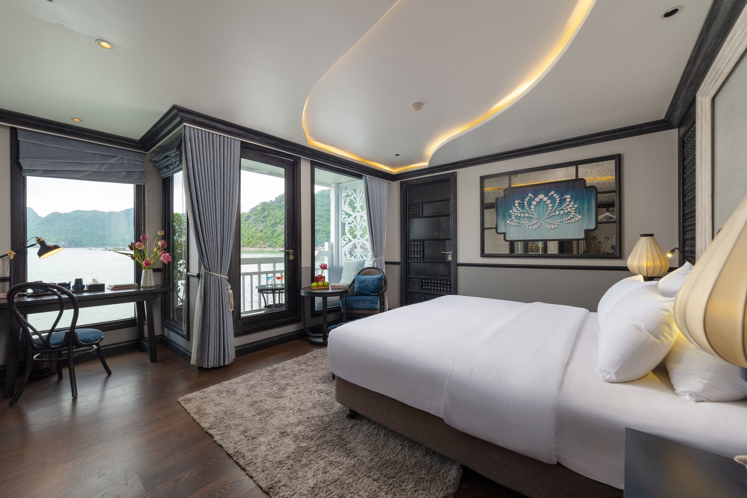 Habitación de hotel moderna con una cama grande, un escritorio con silla, vistas a la montaña y un balcón con mesa y sillas.