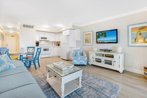 A modern living room with a grey sofa, a glass coffee table, and a dining area with blue chairs.