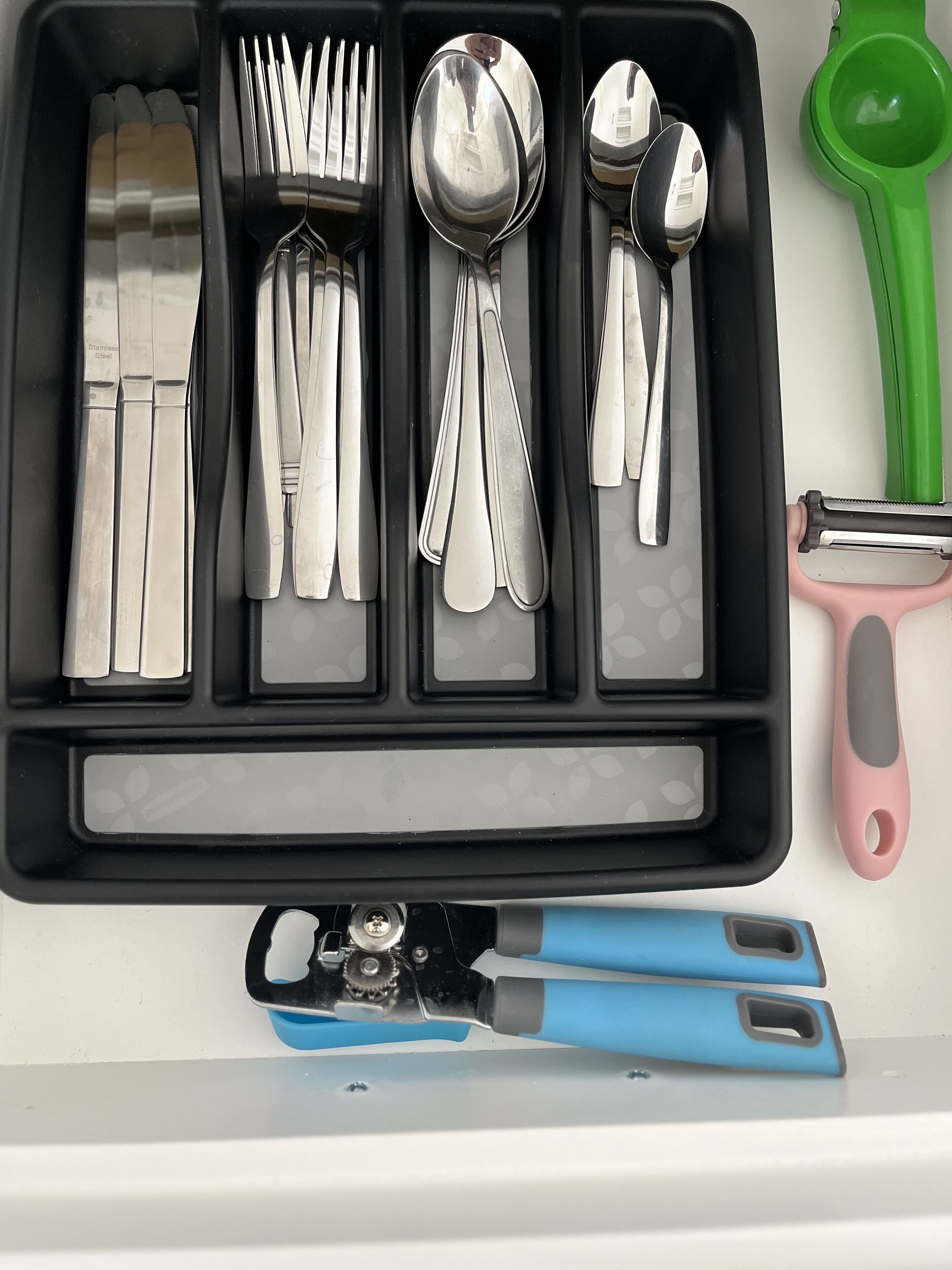 A drawer containing stainless steel cutlery, including forks, knives, and spoons, along with a green and pink peeler.
