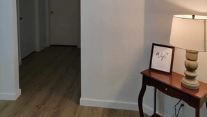 Ein Flur mit Holzboden, einer Tür am Ende und einem kleinen Tisch mit einem gerahmten Bild und einer Lampe.