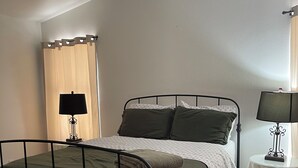 Ein Einzelbett mit Metallgestell, ein Nachttisch mit Lampe und ein Fenster mit Vorhängen.