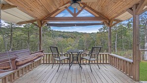 A wooden deck with a covered seating area, a table, and chairs, overlooking a forested landscape. - Cozy Tiny Cabin with a View!! (Decatur)
