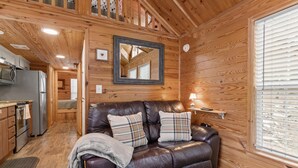 A cozy cabin interior with a leather couch, wooden walls, a kitchen area, and a large mirror. - Cozy Tiny Cabin with a View!! (Decatur)