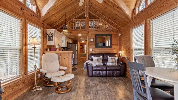A spacious cabin interior with a high ceiling, wooden walls, and large windows. There is a dining area with a table and chairs, a living room with a sofa and a chair, and a kitchen area with cabinets and a countertop. - Cozy Tiny Cabin with a View!! (Decatur)