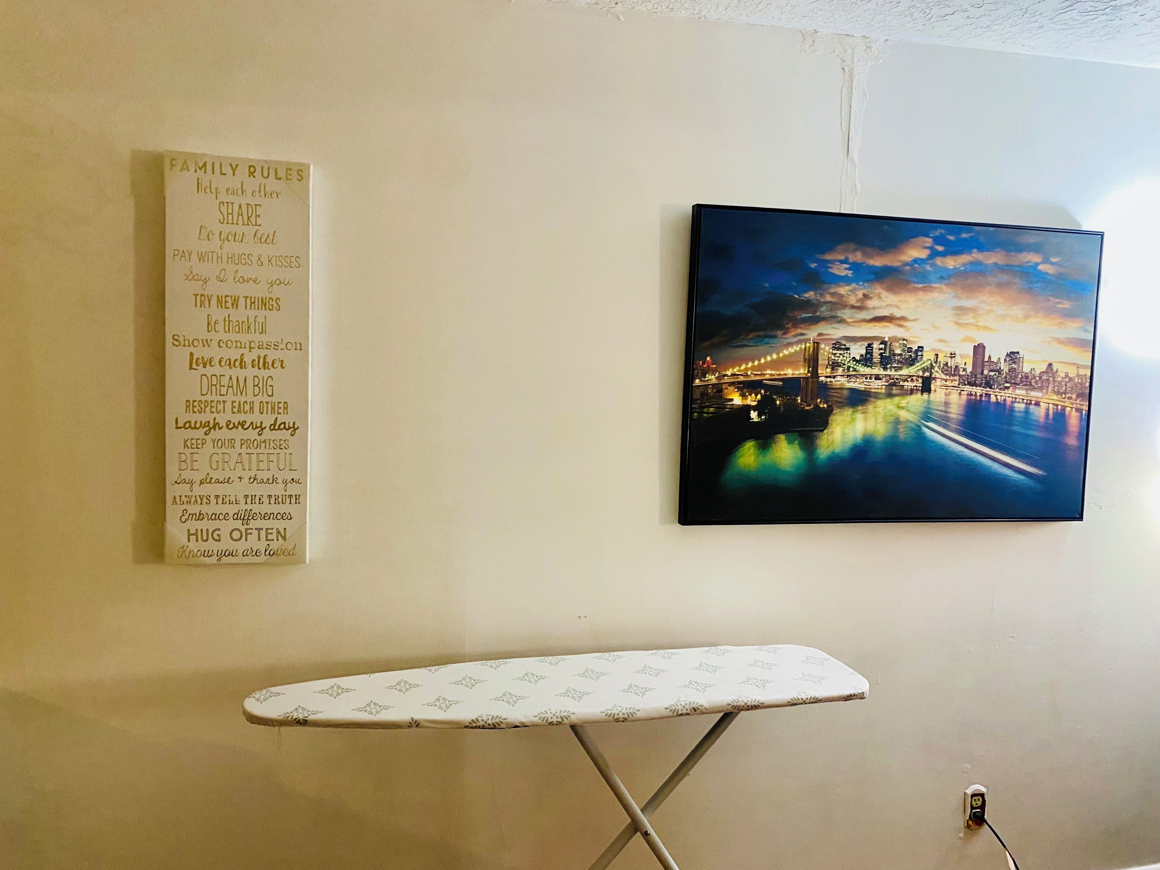 Une pièce dont le mur est orné d’une liste encadrée des règles familiales, avec un tableau représentant un paysage urbain et une planche à repasser au premier plan.