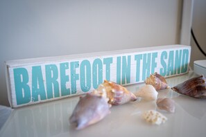 En skylt med texten ”Barfota i sanden” och flera snäckskal på en reflekterande yta.
