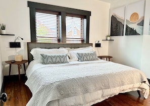 A neatly made bed with a white quilt, two bedside tables with lamps, a window with blinds, and a wall with two framed artworks.