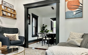 A modern living room with a dark grey sofa, a glass coffee table, and a dining area with a black table and chairs. A large plant is in the dining area, and a painting is on the wall.