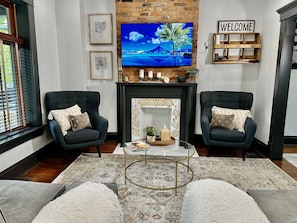 A living room with two dark armchairs, a coffee table, a fireplace, a TV, and a welcome sign.
