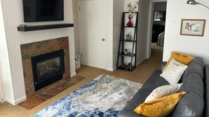 A living room with a fireplace, a television mounted above it, a grey sofa with yellow cushions, and a patterned rug.