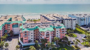 A beachfront hotel with multiple buildings, a parking area, and a beach in the background. - Beach Cottages Ocean & Pool View Condo (Indian Shores)