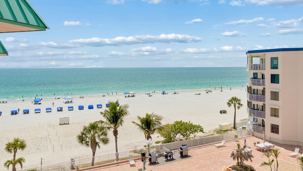 A beach view with a building, palm trees, and lounge chairs. - Beach Cottages Ocean & Pool View Condo (Indian Shores)