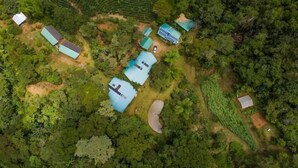 Eine Vogelperspektive zeigt ein Anwesen mit mehreren Gebäuden, darunter ein Haupthaus mit einem großen Dach und kleinere Bauten, umgeben von dichter Vegetation und einem Feld.