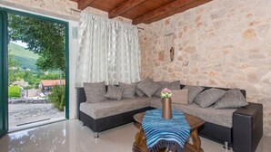 A modern living room with a stone wall, a grey sofa with multiple cushions, a wooden coffee table with a vase of flowers, and a view of a village through a glass door. - Villa Brkic with private pool and playground- ideal for families & friends (Dicmo Osoje)