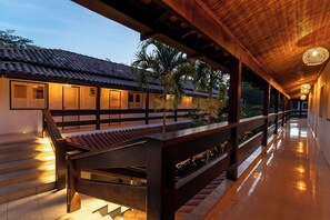 Un paseo techado con barandillas de madera y suelo de baldosas, que conduce a un edificio con tejado de tejas y ventanas iluminadas.