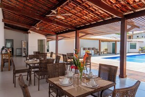 Un área de comedor al aire libre techada con mobiliario de mimbre y una piscina al fondo.