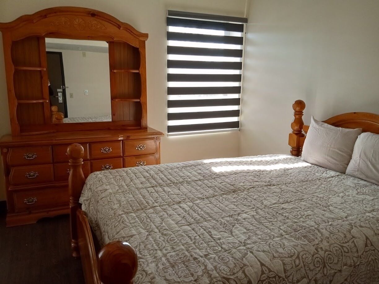 A bedroom with a wooden dresser, a large mirror, a bed with a patterned quilt, and a window with blinds.