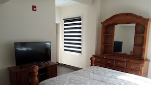 A bedroom with a bed, a wooden dresser, a television, and a window with blinds. - Rochester Hotel (Rochester)