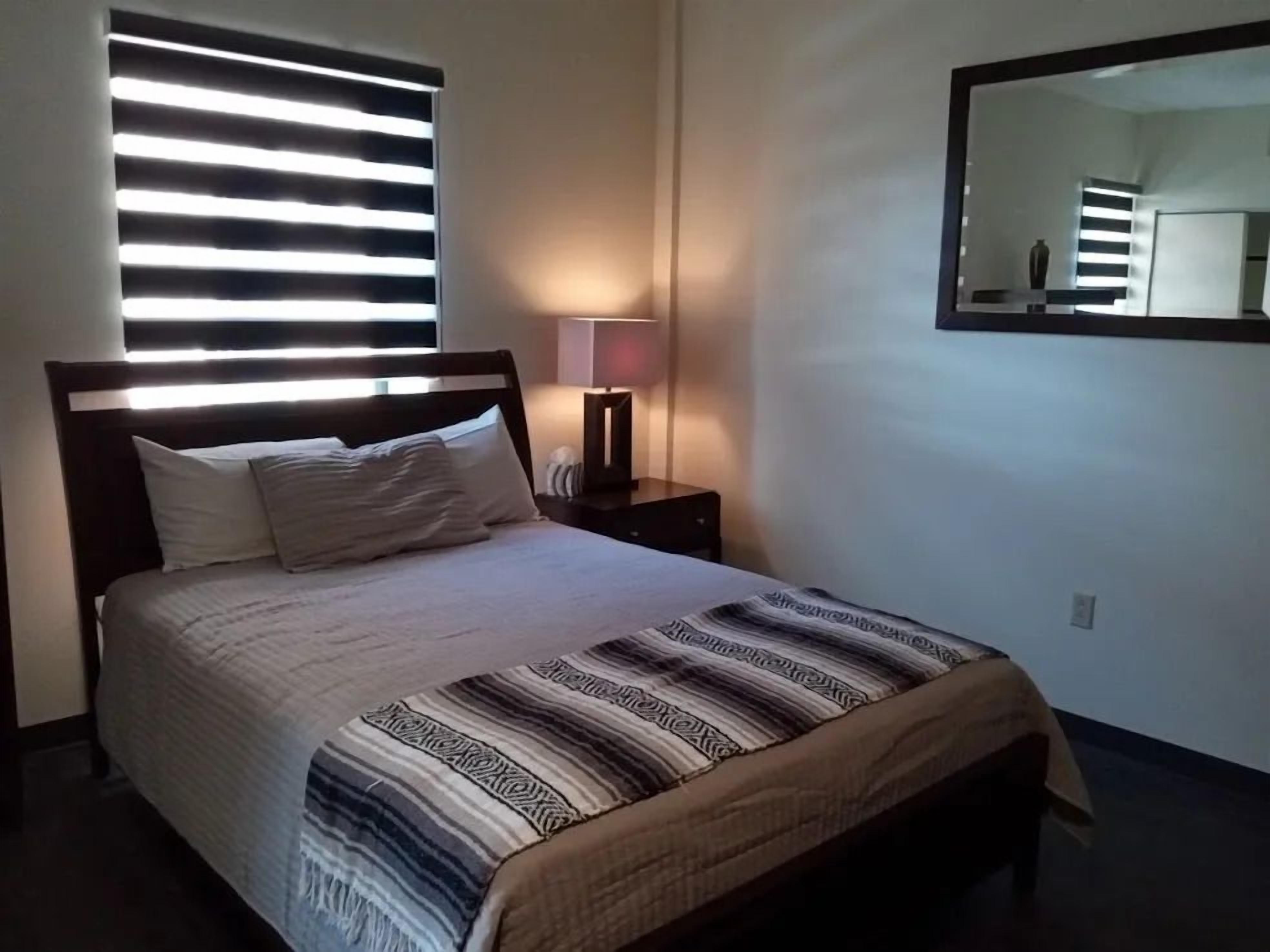 A bedroom with a bed, a bedside lamp, a mirror, and a window with blinds.